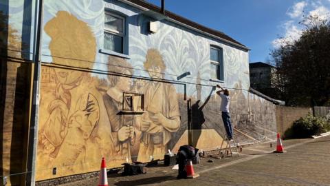 A wall with a painting of a woman a man and a child in mustard next to a painting of a steam engine and railway tracks- a man in a white shirt painting the artwork with spray cans