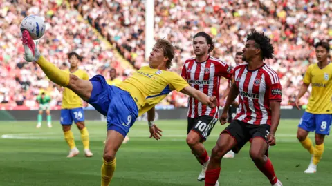 August Priske (left) holds his leg in the air to control the ball. Sheffield United players stand behind him