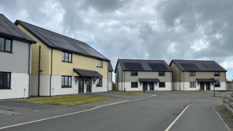 Four semi-detached modern homes at the end of a street. There are solar panels on the roof and wooden cladding on the top of the buildings. Each home is painted a different colour. They are grown, green, yellow and grey.
