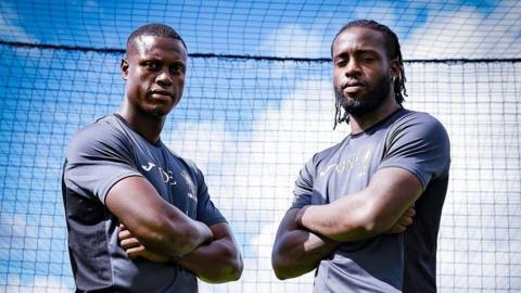 Great and Diop Evans stand next to each other with arms folded and look at the camera. They are both wearing dark blue t-shirts and are standing in front of a net with blue sky behind.