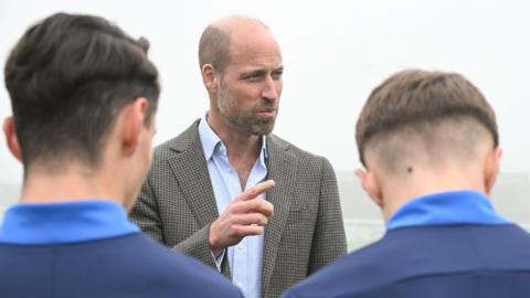 Price William is pointing a finger and two young men are looking at him with their backs to the camera. Prince William is wearing a pale blue shirt and a checked jacket. He has pale grey hair and a beard. It is a foggy day.