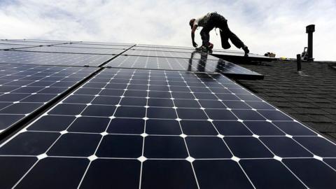 An unidentifiable man silhouetted against a cloudy sky installing solar panels on a roof.