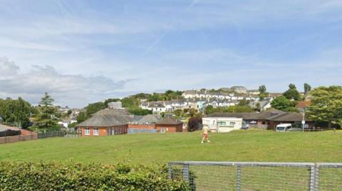 Land for homes off Greenswood Road, Brixham. There is a bush in the front left of the frame and attached to it is a gate. There is a person walking across a field. There are houses in the background. The sky is blue with white clouds. 