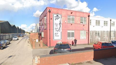 A building with a pink exterior and a red brick wall around its perimeter. It has a large black and white sign that reads "the foxy, dance bar, adult shop, adult cinema". there is a silhouette of a woman leaning against a pole.