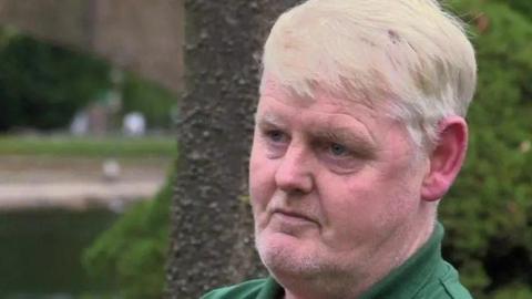 A man in his 50s with sandy hair and a green-collared shirt looks to the side of the camera.