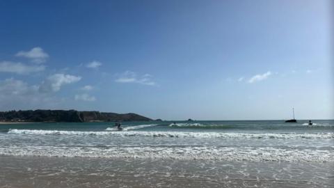 People in the water at St Brelade Bay. It is a sandy beach and there's a view of waves crashing on the sand. There's land in the distance on the left. The sky is blue with a few white clouds.