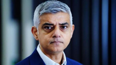 A close shot of Sir Sadiq, wearing a blue suit, looking into the camera with a neutral expression