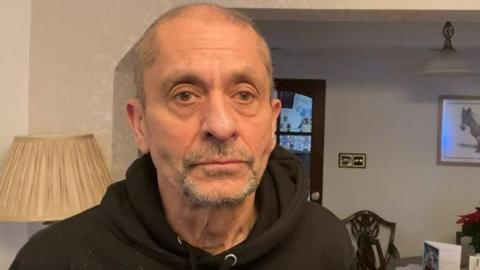 An older man with a short, grey beard looks unsmiling into the camera. Behind him is a doorway of glass with brown, wooden edges and a lamp with a beige lampshade on it. A picture hangs on the wall showing the head of a donkey in shot.