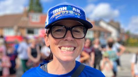 A woman smiles at the camera. She is wearing a blue cap and a blue T shirt and big black-rimmed glasses