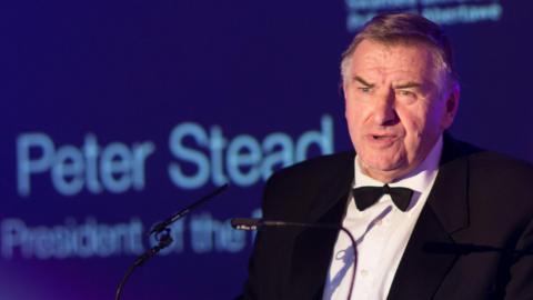 Prof Peter Stead speaking into a microphone wearing a black dinner jacket, white shirt and black tie, with the beginning of the sentence Peter Stead President of in the blurred blue background behind him.