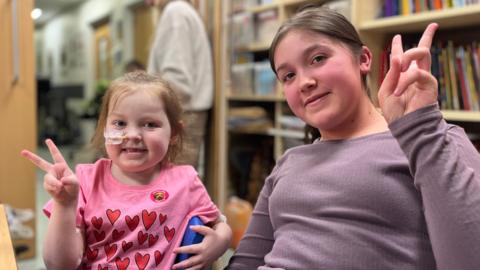 Sisters Arne and Grante sat next to each other with peace signs. They are smiling at the camera sat in the school facility in the hospital.