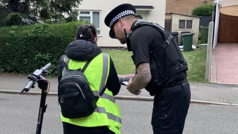 A police officer stands next to a woman seen from behind, as they discuss her e-scooter. She is wearing a high-vis neon vest and grey jogging trousers.