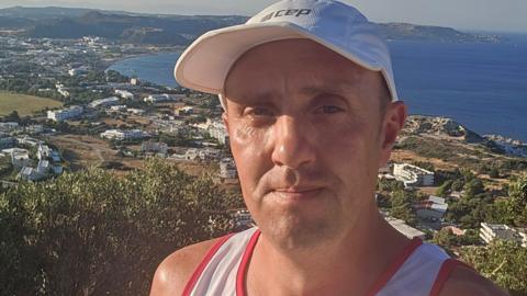 A man in his 40s stood against the backdrop of a coastal setting. He is wearing a white baseball cap and vest top. Behind him is the sea and a smattering of white properties.