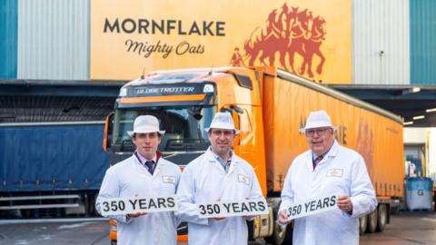 Three men in white overalls hold numberplates which read '350 years' while stood in front of a large industrial unit with Mornflake branding and a yellow HGV with the same branding.