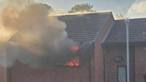 The upper floor of a brick residential building. A fire is blazing inside a window and thick smoke is billowing out.