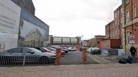 A small, open, ground-level car park with a three-storey red brick hotel on one side and a large property advertisement on the other. A metal fence runs across the entrance-side, bordered by two brick columns.