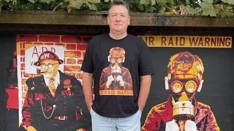 Kevin Preston stands in front of his two murals of the air raid warden and a boy in a gas mask. He has short brown and grey hair and is wearing light blue shorts and a black T-shirt with the image of the boy in a gas mask on it. 