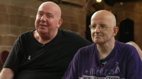 Graham and Ian Hopkins sitting next to each other. Graham is wearing a black top and Ian is wearing glasses and a purple top