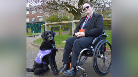 A woman sits in a wheelchair and smiles. She holds the lead of a black Labrador, which wears a purple vest.