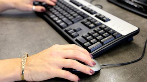 A person's hands with dark nail varnish and a gold bracelet using a mouse and computer keyboard