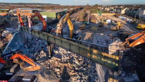 Diggers and other construction machinery are surrounded by rubble as a bridge is demolished over a railway track. It is a sunny winters day in a residential area.