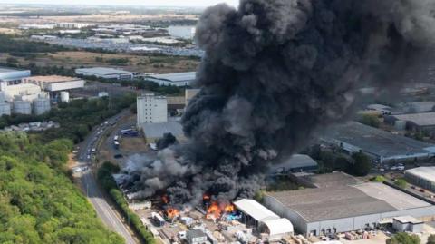 A large fire at an industrial estate, there are flames and a large thick plume of black smoke coming out of the building. There are cars parked nearby. It is an overheard view of the site.