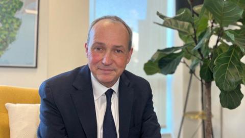 Lyndon Farnham pictured in a navy blue suit, with white shirt and navy blue tie. His hair is short and grey. He's sitting on a yellow chair with a plant visible in the background of his office. He is smiling.