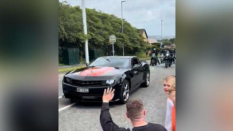 A black supercar leading the special vehicle parade in Bury followed by a number of motorbikes on a main road. It is a dry day and hills and the countryside can be seen in the background.