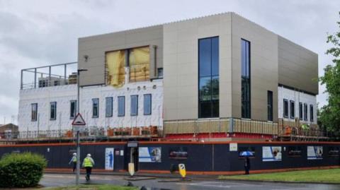 A two and three storey flat-roofed building under construction in Peterborough. It is surrounded on the ground floor by a tall blue barrier. A man in a hi-vis yellow jacket and white helmet is crossing a road towards the construction site. 