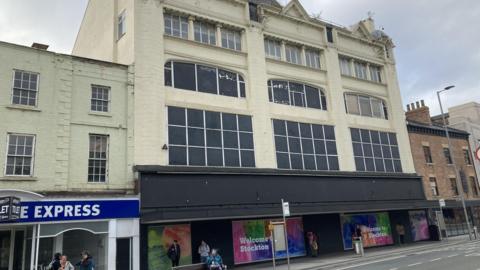 View of the former Debenham's store in Stockton. The cream coloured building, dating from the 1890s has high windows on three levels. On the ground floor are big display windows with posters stating "Welcome to Stockton".
