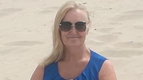 A woman with long blonde hair, sunglasses and a blue sleeveless top, smiles as she stands on a beach.