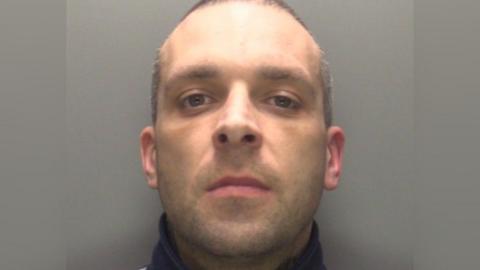 Police custody shot of a short-haired man who has dark eyes and stubble and is looking straight at the camera