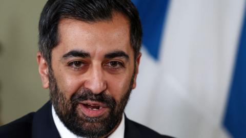 Humza Yousaf, with dark hair and a dark beard, photographed in a close-up shot with this mouth open as he speaks. A saltire is hanging on a wall behind him. 