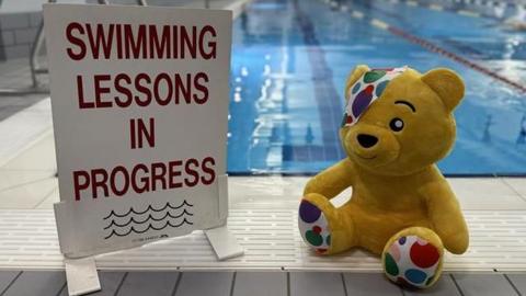 Plushie of Pudsey the bear, in front of a pool, next to a sign which says 'Swimming lessons in progress'.