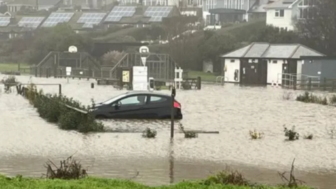 A black car in brown flood water up to the wheel arches. There is no one in the vehicle. A flooded play park can be seen in the background.