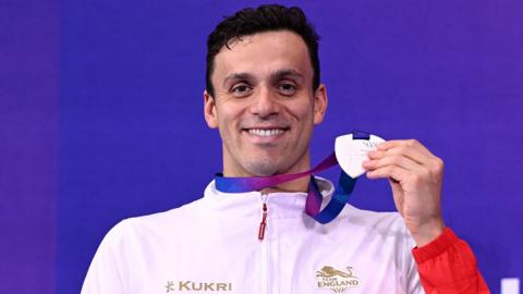 James Guy, wearing England's white and red swimming training jacket, smiles as he holds up his Commonwealth Games silver medal for the men's 200m freestyle