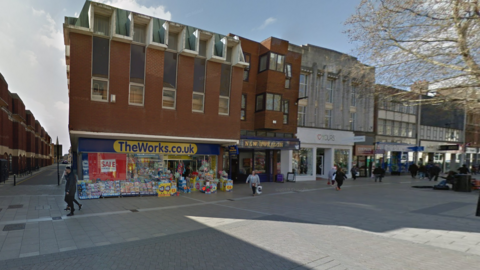 A pedestrianised street with a red-brick building with a sign above the entrance that reads: "The Works". The next door unit has a blue frontage with a sign that reads "New Horizon". 