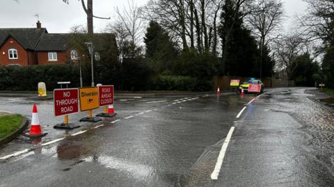 A street covered in water. Multiple roads signs are up marking a road closure and the diversion route. A police car can be seen in the background.