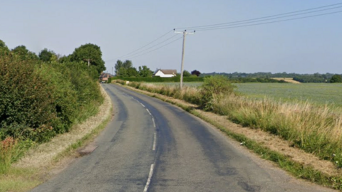 A Google Maps street view image of a B-road flanked by farmland and green bushes and trees. A white house can be seen in the distance. 