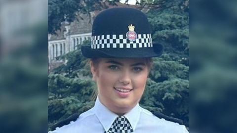 Hannah Byrne looks at the camera in her policing uniform. She is wearing a hat with the Surrey Police logo on it and a black and white checked neckerchief. 