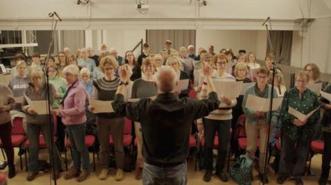 Sing Out! South Craven Choir are pictured looking at their song sheets while a conductor stands in front of them