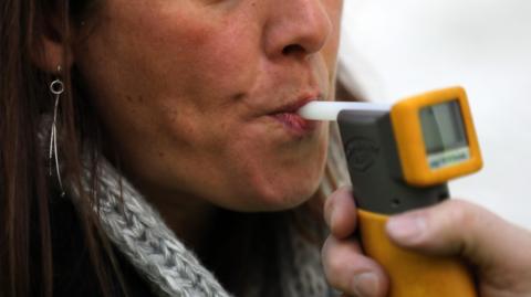 The partially-obscured face of a woman with long brown hair, silver earrings and a grey scarf blowing into an electronic breathalyser machine made of yellow and grey plastic with a white breathing tube. 