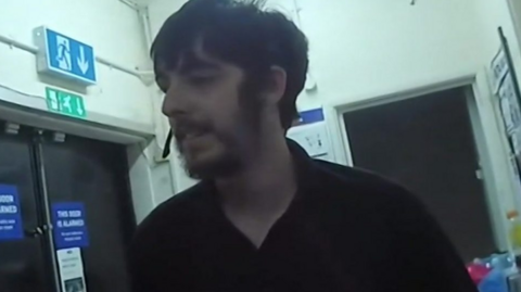 Man in corridor with fire doors behind him - wearing a head microphone. He has a black top on and dark hair and a dark beard