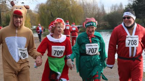 Four people dressed in festive attire, holding hands and running along Bedford's Embankment. The man on the left is wearing a reindeer onesie, a woman is wearing a Mrs Santa dress, an elf is next to her, and on the right is a man dressed in a Santa suit with a black hat. They all have numbers on them. Other runners in Santa suits, a river, a path and trees are behind them 