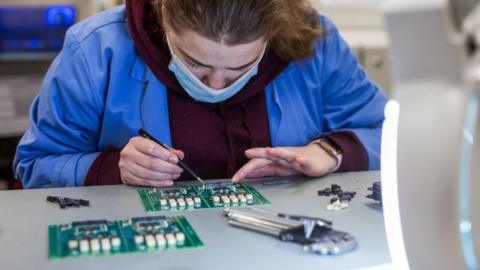 This image captures the careful and precise work of someone assembling or repairing electronic circuit boards. The person is using a tool—possibly a soldering iron or tweezers—to manipulate small components on the board. The workspace is well-lit, suggesting a focus on detail and accuracy. Various circuit boards, electronic components, and tools are laid out on the table, emphasizing the complexity and skill involved in electronics work.