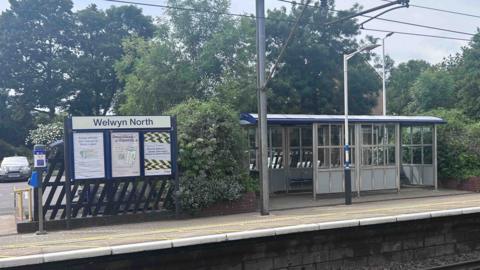 Welwyn North railway station, with a shelter to the left, railway tracks in front, a platform and trees behind the shelter. A sign is to the left. 
