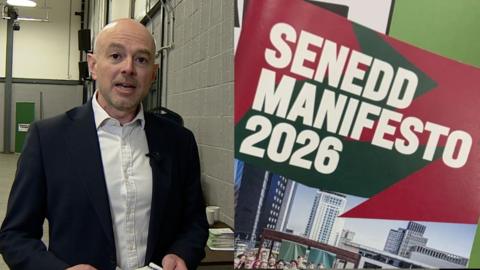 The image is split into two halves. On the right hand side there is a man wearing a dark blue suit jacket with a white shirt that has the top button undone. The background consists of white and gray walls and they are covered with pipes, there is also a table with coffee mugs behind him. On the right hand side of the image there is a Wales Green Party manifesto that reads SENEDD MANIFESTO 2026 on the cover and is a mixture of red and green, there are also skyscrapers and a large group of people on this front page. 