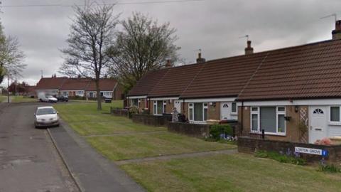 A row of bungalows on the right hand side at the entrance to the road, with more bungalows seen in the distance. There is grass in front of the houses and the occasional tree.