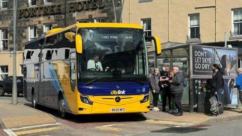 Bus buidhe agus gorm le daoine a' feitheamh airson a dhol air bòrd.