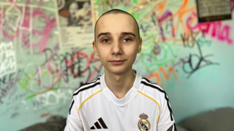 Leo sits on his bed in his bedroom, behind him is a colourful graffiti wall. He is wearing a signed Real Madrid top which was gifted to him by Jude Bellingham. He has a buzz cut.